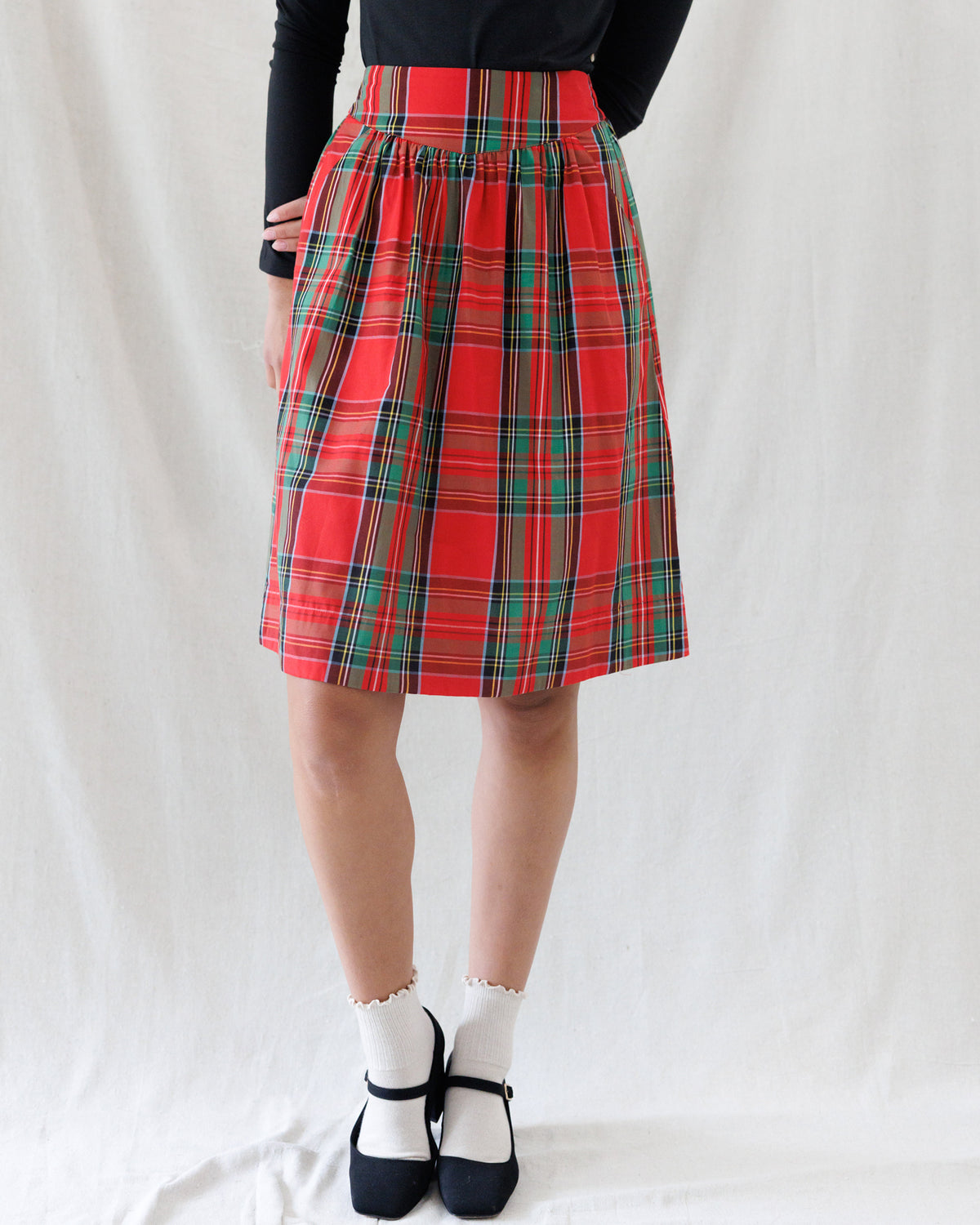 A red and green tartan plaid pleated skirt worn by a model, paired with a black long-sleeve top, white socks, and black Mary Jane shoes, photographed against a white fabric backdrop.
