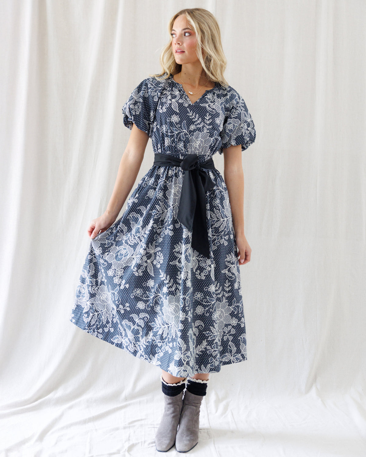 A woman modeling a black and white floral print midi dress with a black waist tie, paired with gray ankle boots, standing against a white draped fabric backdrop.