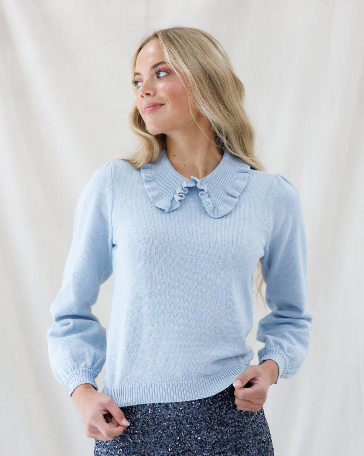 A woman wearing a light blue knit sweater with a ruffled Peter Pan collar and a matching bow at the center of the collar, paired with a dark sparkly skirt, posed against a draped white fabric background.