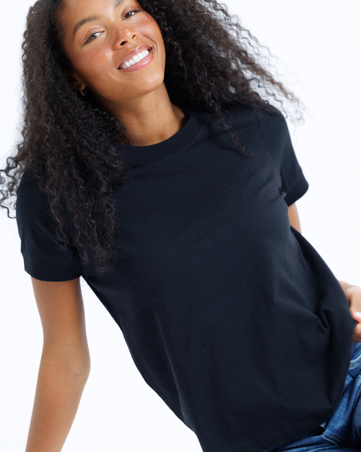 A smiling woman with long curly black hair wearing a plain black short-sleeve crewneck t-shirt and blue jeans, photographed against a clean white background.
