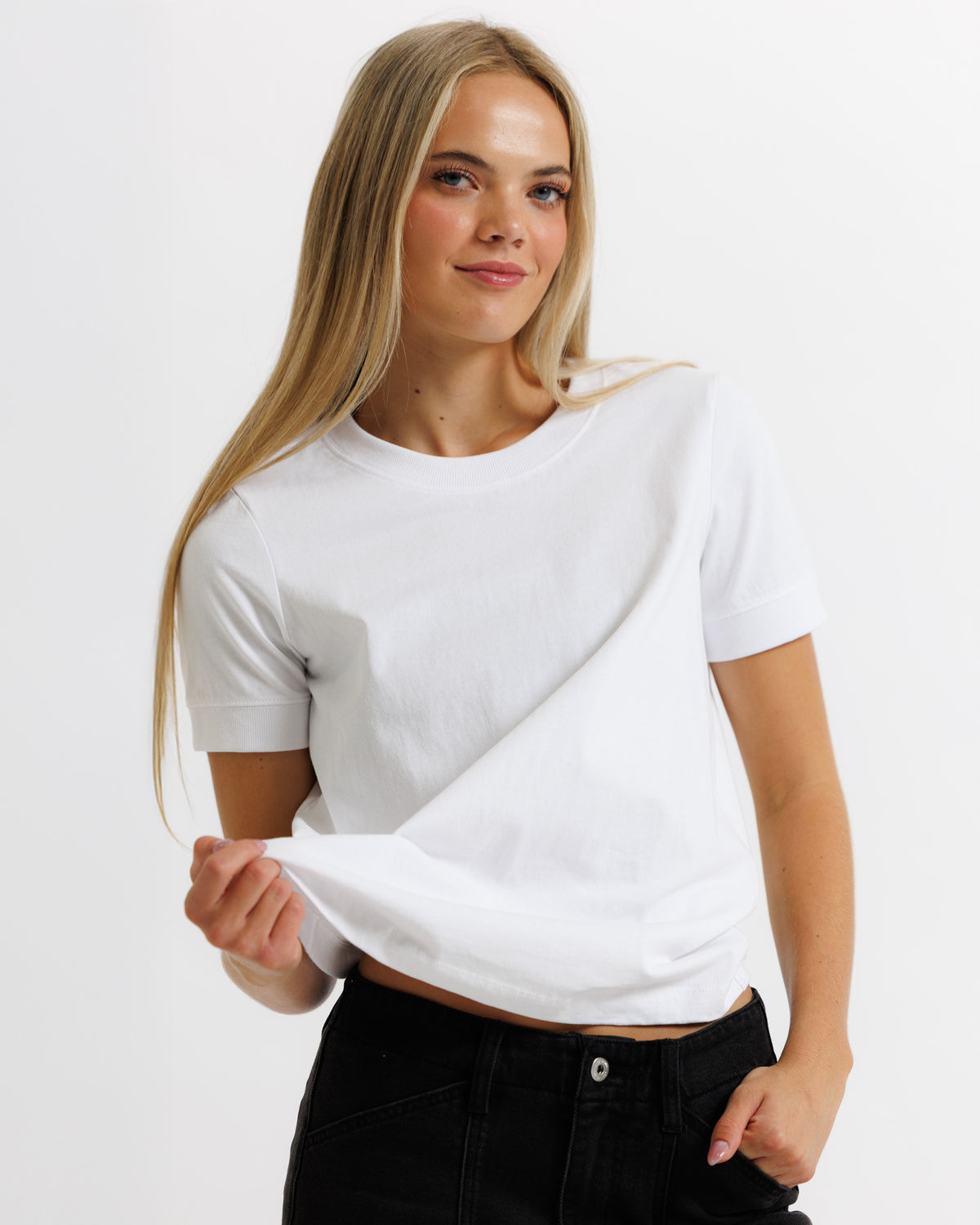 A woman with long blonde hair wearing a white short-sleeve crewneck t-shirt and black jeans, posing against a solid white background.