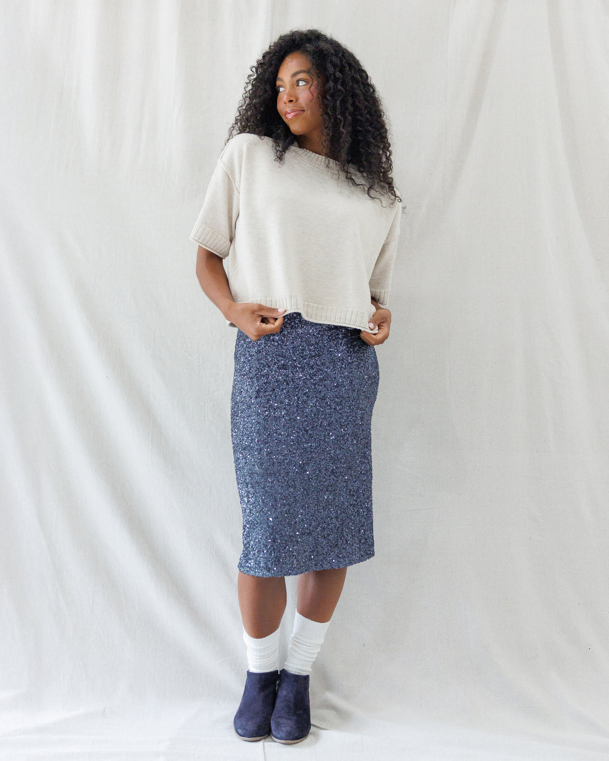 Model wearing a boxy beige short-sleeve knit sweater with a ribbed crew neckline and wide hem, styled over a sparkly navy sequin midi skirt.