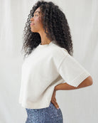 A woman with long, curly black hair is shown in profile wearing a cream-colored short-sleeve knit sweater and blue sequined pants against a white studio backdrop.