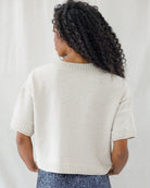 Back view highlighting the clean, loose fit of the beige knit top, detailed with exposed drop-shoulder seams, a thick ribbed collar, and matching ribbed hem