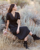 A woman wearing a black midi dress with short puff sleeves and a lace-trimmed V-neck sits on a rock in an outdoor desert landscape with dry grass and shrubs.
