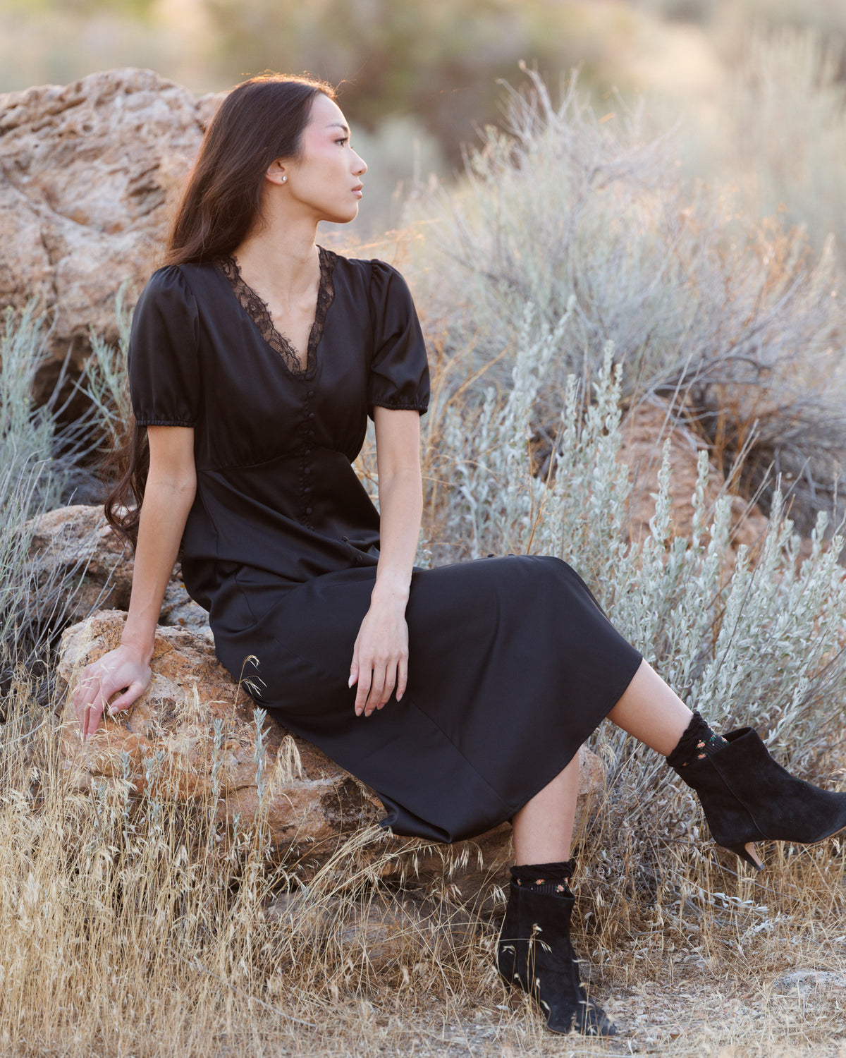 A woman wearing a black midi dress with short puff sleeves and a lace-trimmed V-neck sits on a rock in an outdoor desert landscape with dry grass and shrubs.