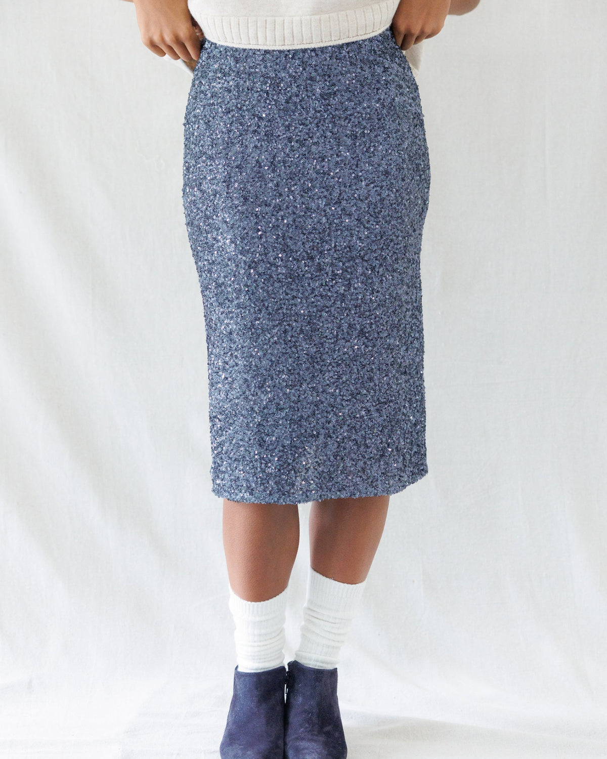 A person wearing a textured blue and white speckled knit pencil skirt, paired with a cream sweater, white socks, and navy blue slip-on shoes, standing against a white fabric backdrop.