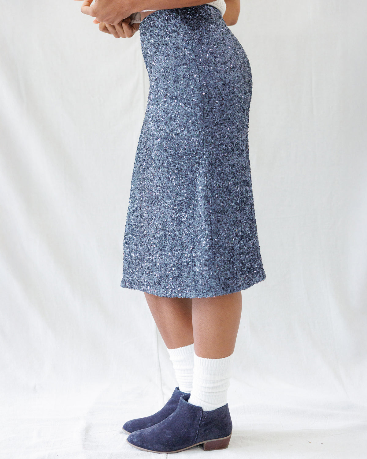 A person wearing a knee-length blue sequined skirt, paired with white socks and navy blue suede ankle boots, standing against a white fabric backdrop.
