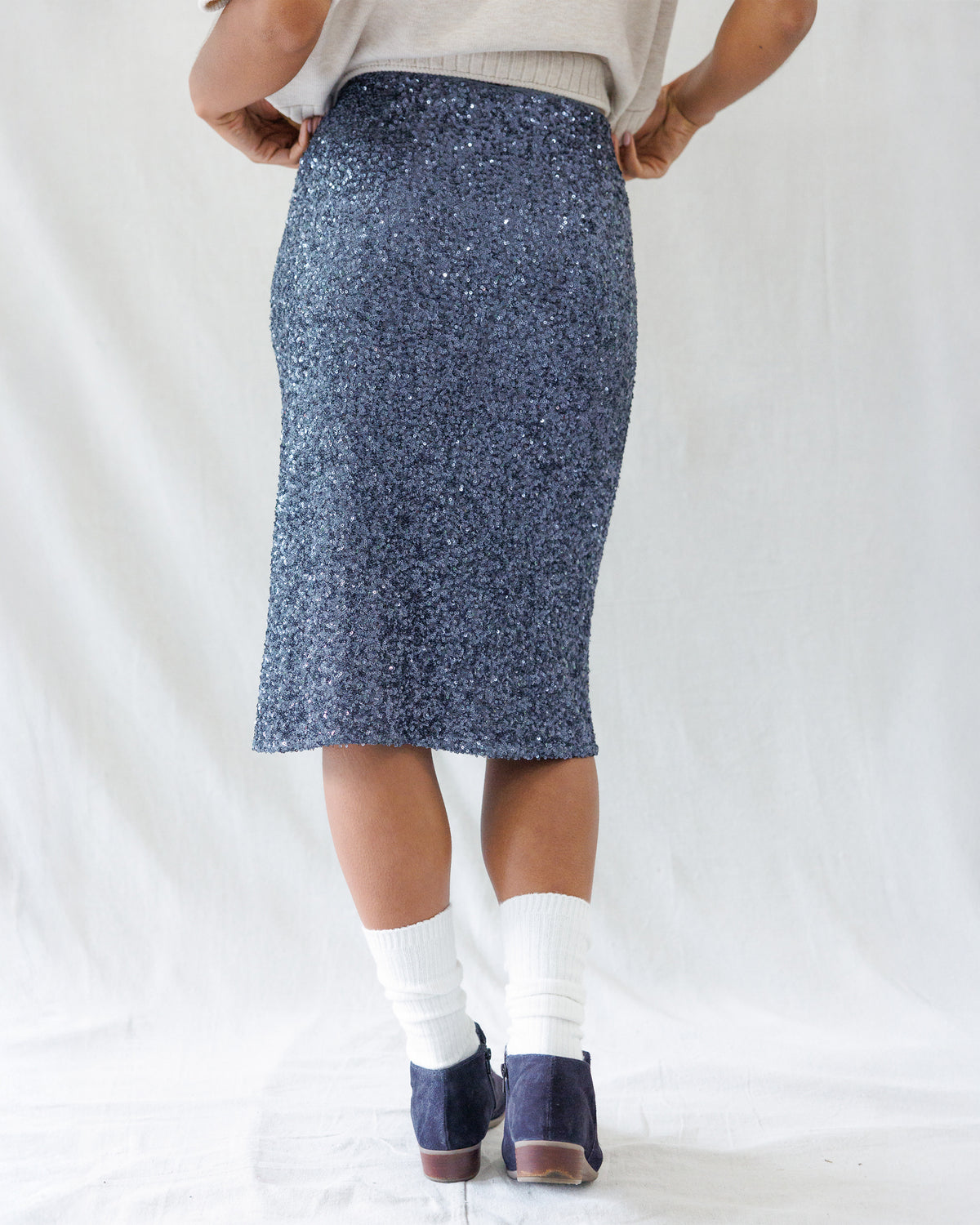 A person shown from the back wearing a knee-length navy blue sequined skirt, a light beige top, white socks, and navy shoes against a white fabric backdrop.