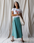 Woman wearing a white t-shirt with strawberry design and a green skirt, holding a beige tote bag against a plain background.