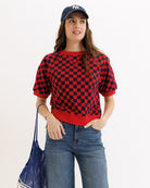 Person wearing a red and black checkered sweater with a blue cap and jeans, holding a blue bag on a white background.