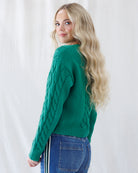 A side view of a woman wearing a green cable-knit sweater and blue jeans, standing against a white curtain background.