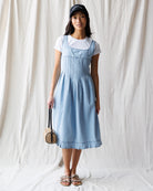 Woman wearing a light blue dress over a white shirt, holding a woven bag against a plain background