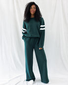 A woman modeling a dark green two-piece lounge set consisting of a long-sleeve sweatshirt with white stripes on the sleeves and matching wide-leg sweatpants, set against a white draped fabric background.