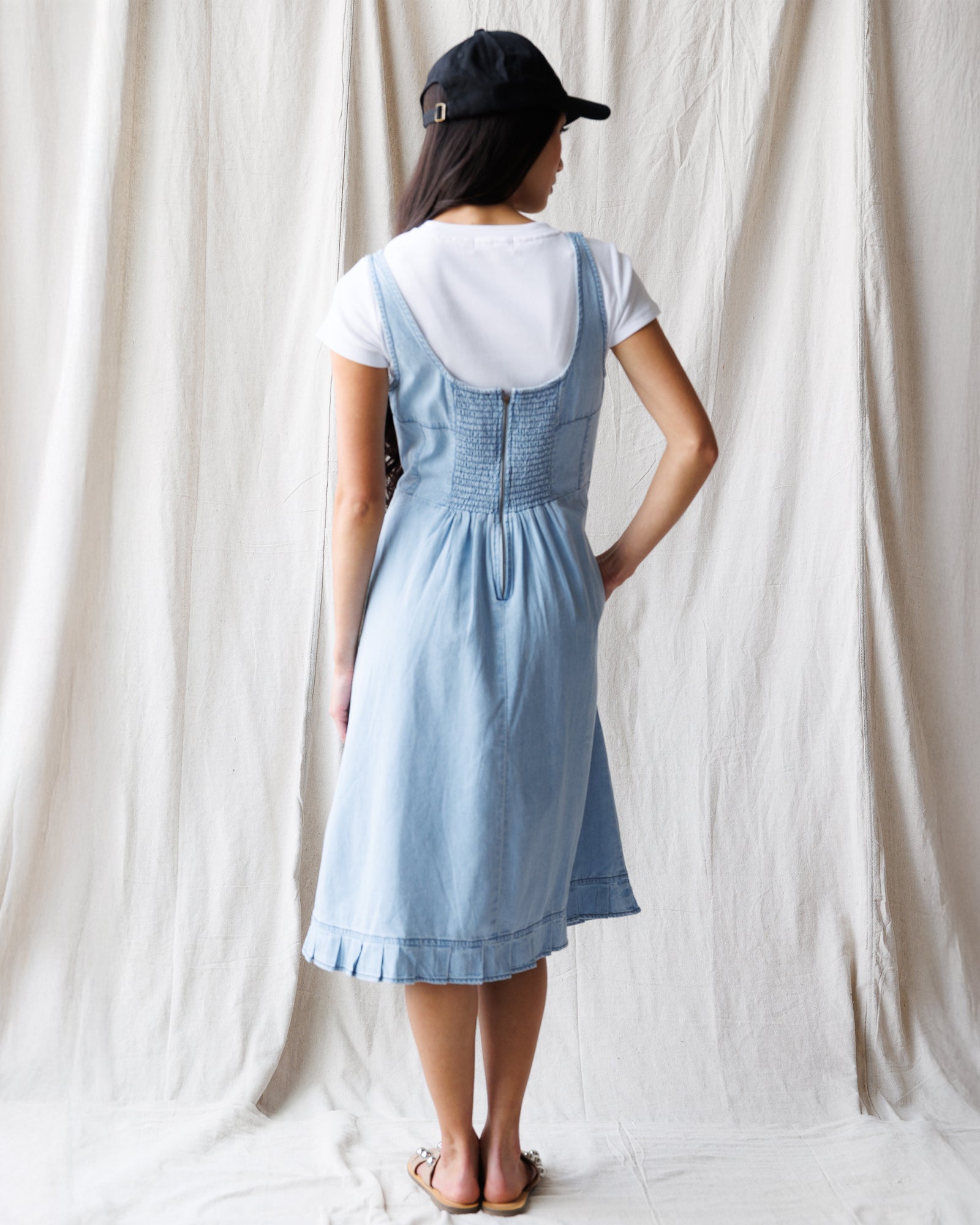 Person wearing a blue denim dress over a white shirt against a white curtain background