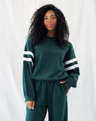 A woman with long, curly black hair wearing a dark green sweatshirt and matching sweatpants with white stripes on the sleeves, standing against a white fabric backdrop.