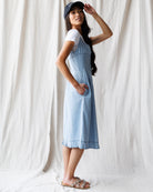 Woman wearing a light blue dress over a white shirt with a black cap, standing against a white curtain.