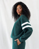 A woman with curly black hair wearing a dark green sweatshirt and matching sweatpants, featuring white athletic stripes on the sleeves, posed against a white draped fabric background.