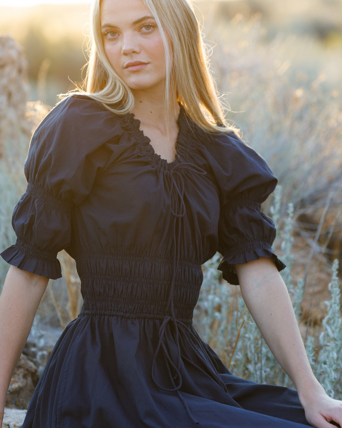 A woman wearing a black puff-sleeve dress with a smocked waist and drawstring tie, posed outdoors in a sunlit field of dry grass.