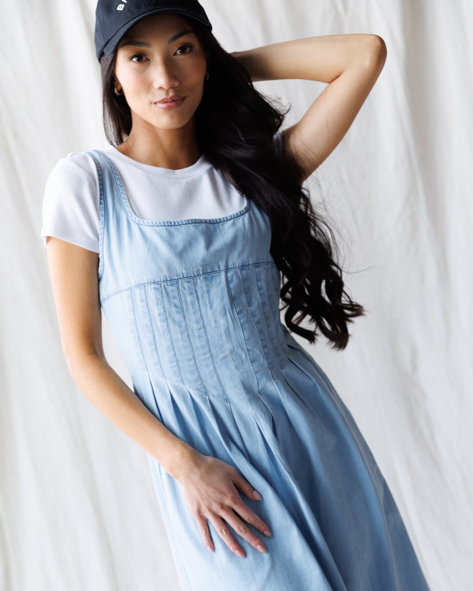 Woman wearing a light blue denim dress over a white t-shirt with a navy cap on a plain background