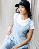 Woman wearing a light blue denim pinafore dress over a white t-shirt with a black cap.
