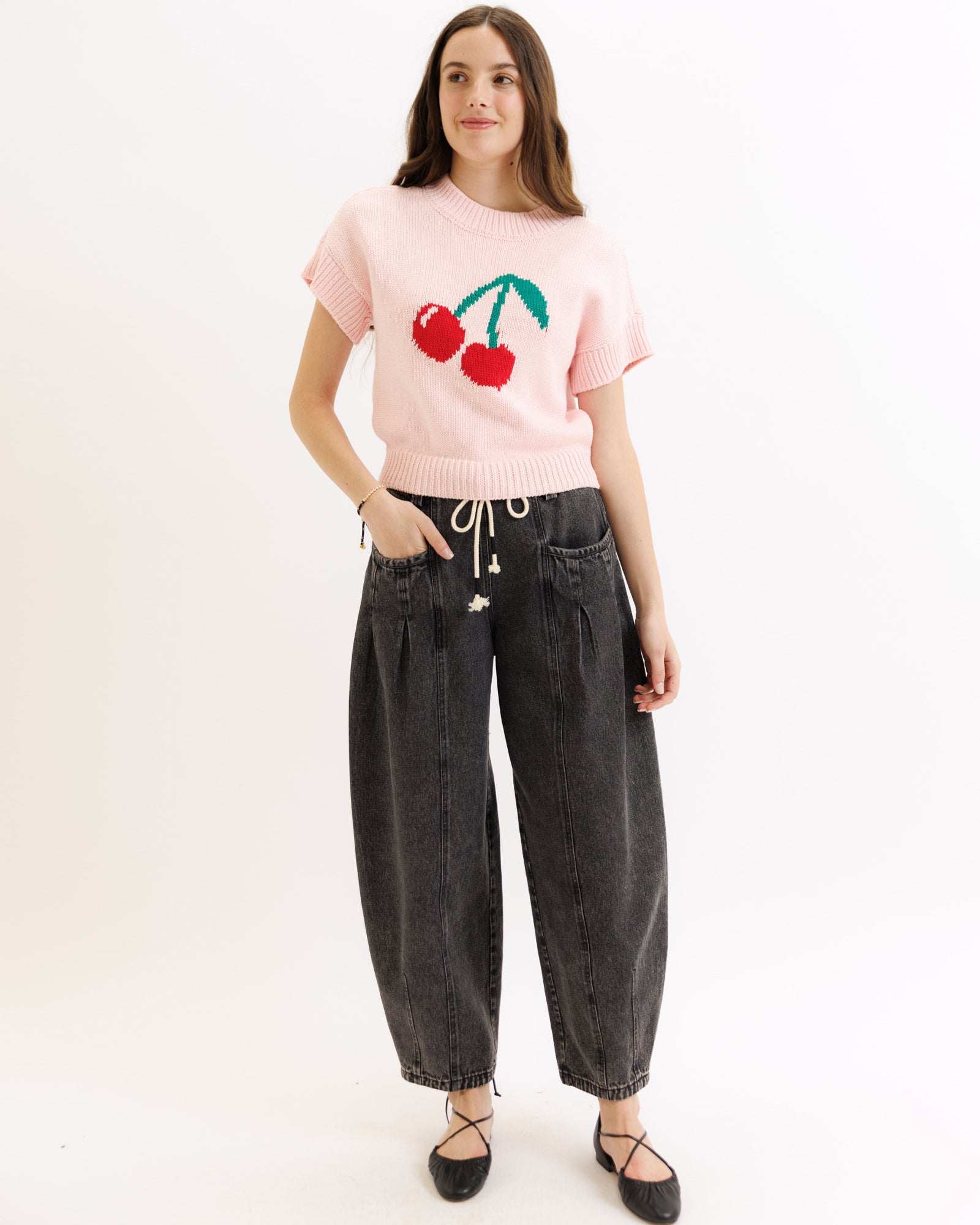 Person wearing a pink sweater with cherry design and black jeans on a white background