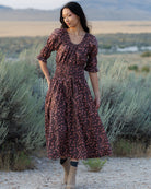 A woman wearing a dark brown midi dress with a small pink floral pattern stands in a desert landscape with sagebrush and mountains in the background.