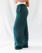 A person wearing dark teal wide-leg sweatpants with an elastic waistband, shown from the waist down against a white fabric backdrop.