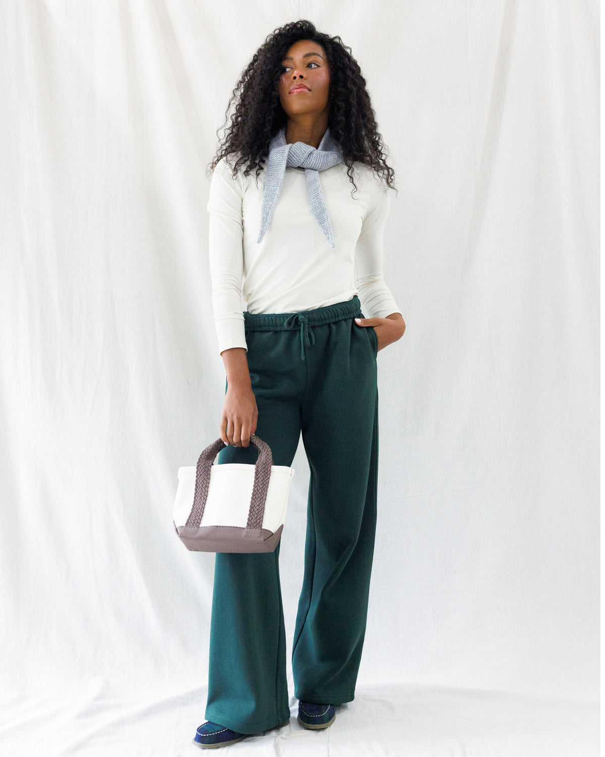 A model stands against a white draped fabric backdrop wearing a white long-sleeve top with a light blue neck scarf, dark green wide-leg trousers, and holding a white and taupe tote bag.