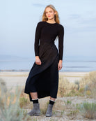 A woman stands outdoors in a dry, grassy landscape wearing a black long-sleeve midi dress with a pleated skirt and gray suede ankle boots.
