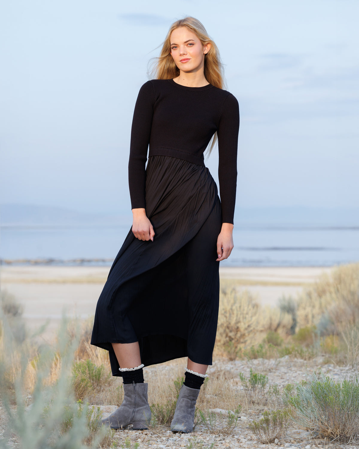 A woman stands outdoors in a dry, grassy landscape wearing a black long-sleeve midi dress with a pleated skirt and gray suede ankle boots.