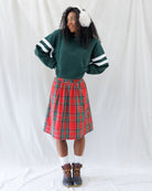 A woman stands against a white fabric backdrop wearing a dark green crewneck sweatshirt with white stripes on the sleeves, a red and green plaid pleated skirt, white socks, and brown boots. She is also wearing white faux-fur earmuffs.