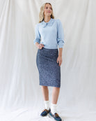 A woman stands against a white fabric backdrop wearing a light blue long-sleeve blouse with a ruffled Peter Pan collar and a dark navy textured pencil skirt. She is wearing white socks and blue sneakers.