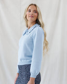 A blonde woman wearing a light blue long-sleeve knit sweater with a ruffled collar and dark patterned pants, standing against a draped white fabric backdrop.