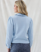 Back view of a blonde woman wearing a light blue long-sleeve knit sweater with a ruffled collar and dark patterned pants, standing against a draped white fabric backdrop.