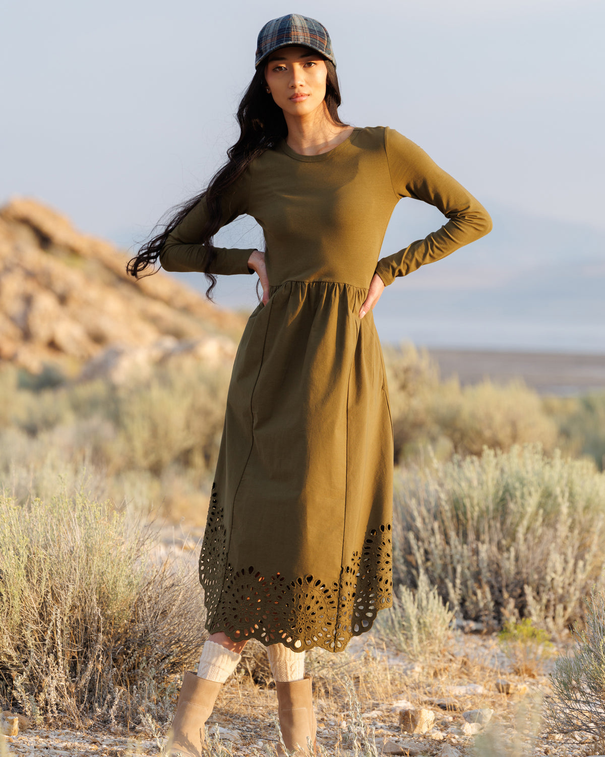 A woman stands outdoors in a desert landscape wearing a long-sleeve olive green midi dress with a gathered waist and decorative cutout hem, along with a plaid cap and beige boots.