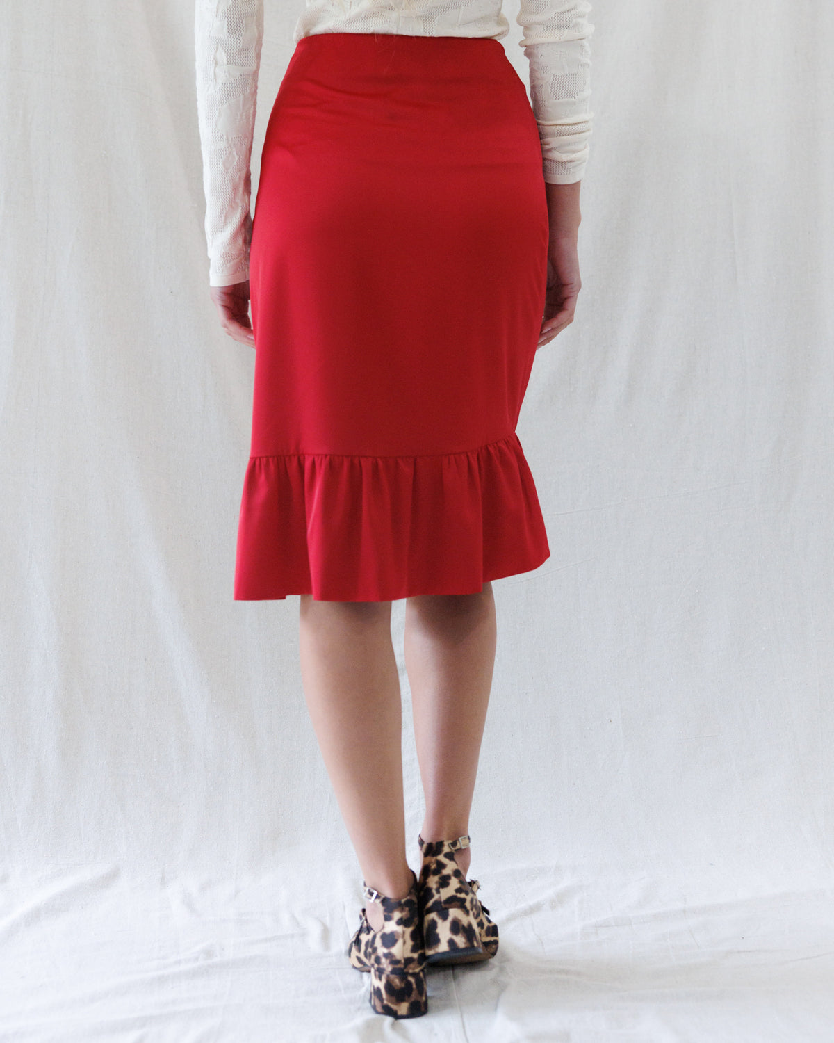 A person shown from the waist down wearing a bright red knee-length skirt with a ruffled hem, paired with a white long-sleeve top and leopard-print ankle boots, standing against a white fabric backdrop.