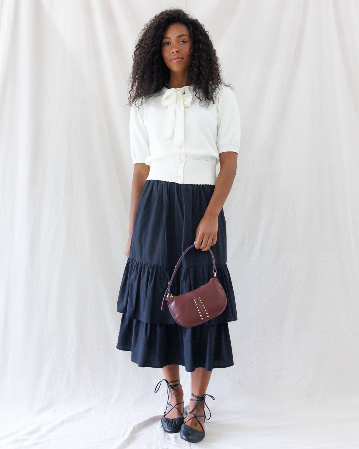 Model wearing a black tiered midi skirt styled smoothly at the waistband under a cream bow-tie short-sleeve cardigan.