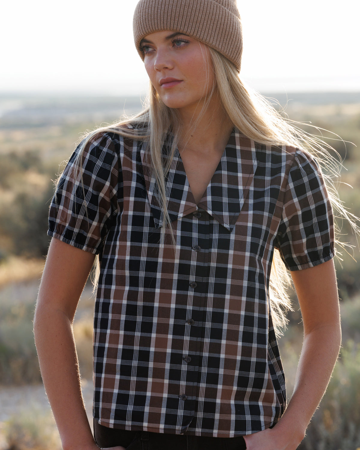 A woman wearing a brown, black, and white plaid short-sleeve button-up shirt and a beige knit beanie, standing outdoors in a sunny, dry landscape.