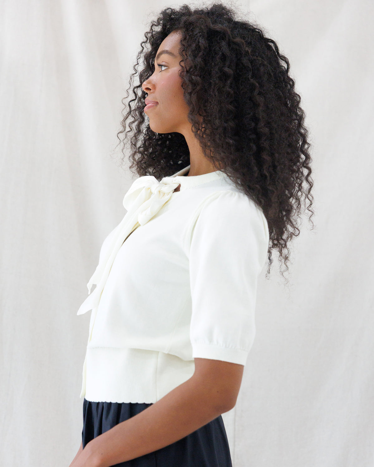 Side profile showing the tailored fit of the cream short-sleeve cardigan, featuring gathered puff sleeves, a statement bow tie neckline, and a wide ribbed waistband.