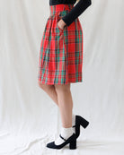 A person wearing a red tartan plaid pleated skirt with green and black stripes, paired with a black long-sleeve top, white socks, and black Mary Jane heels, standing against a white fabric backdrop.