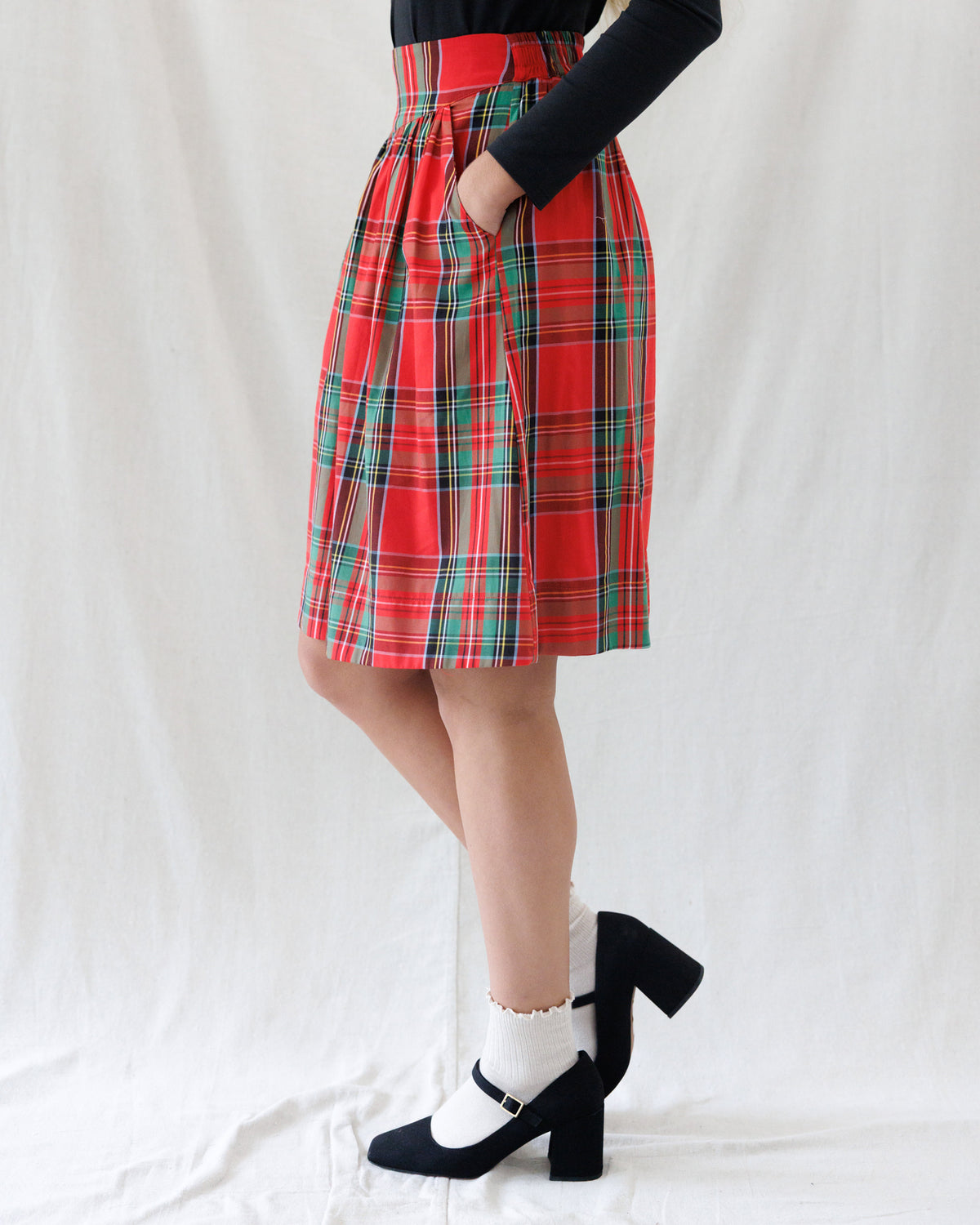 A person wearing a red tartan plaid pleated skirt with green and black stripes, paired with a black long-sleeve top, white socks, and black Mary Jane heels, standing against a white fabric backdrop.