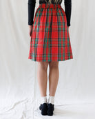 A person shown from the back wearing a red tartan plaid skirt with green and black stripes, paired with a black long-sleeve top, white socks, and black shoes, standing against a white fabric backdrop.