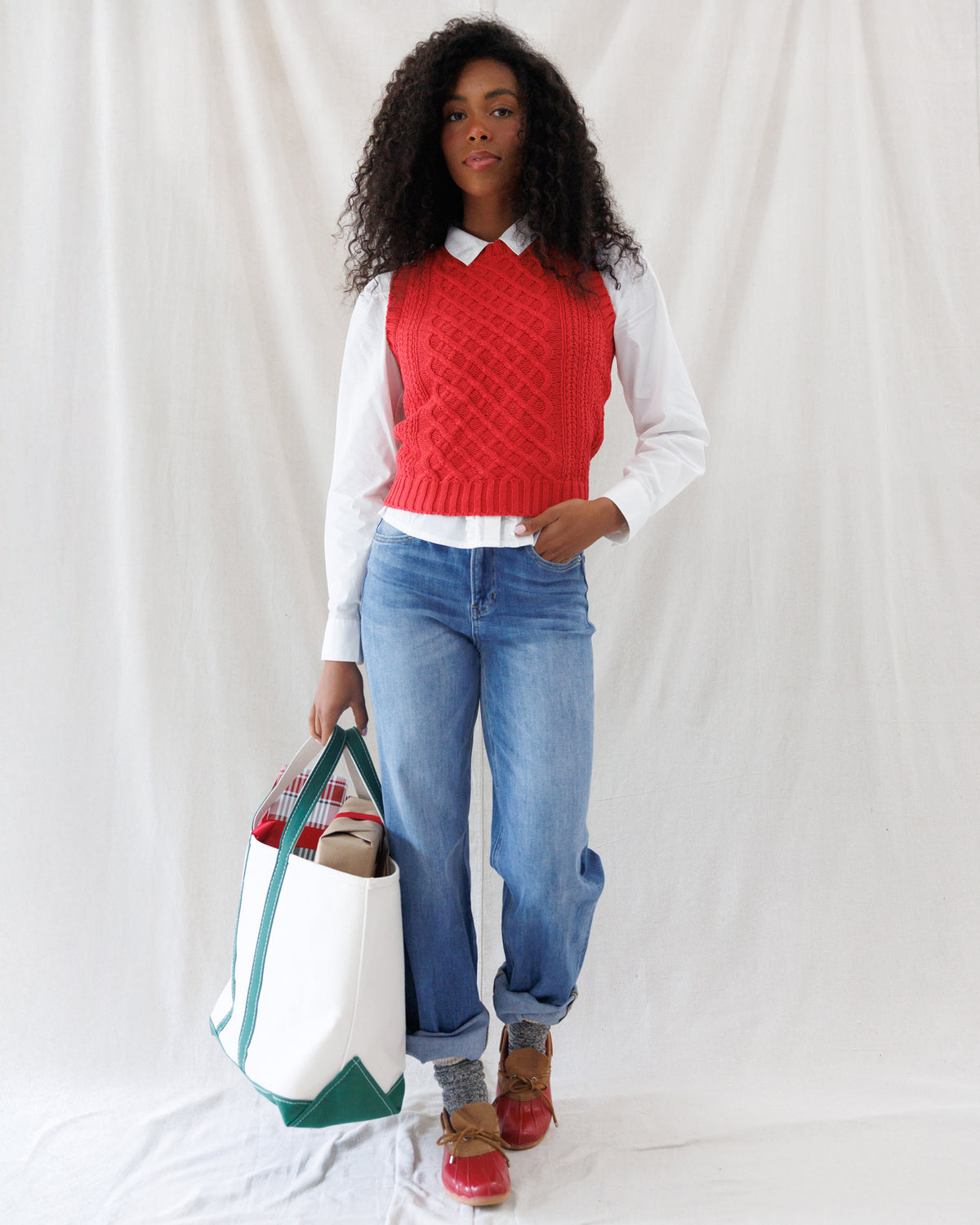 Model wearing a bright red cable-knit sweater vest styled over an untucked white collared button-down shirt with relaxed-fit blue jeans.