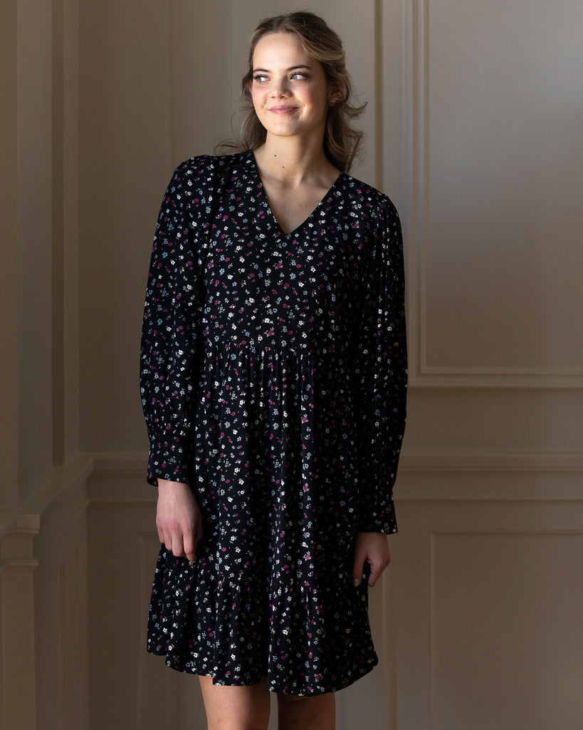 Model wearing a black dress with grey, white and red dainty floral print, with long sleeves, a v-neck and tiers that is knee length.