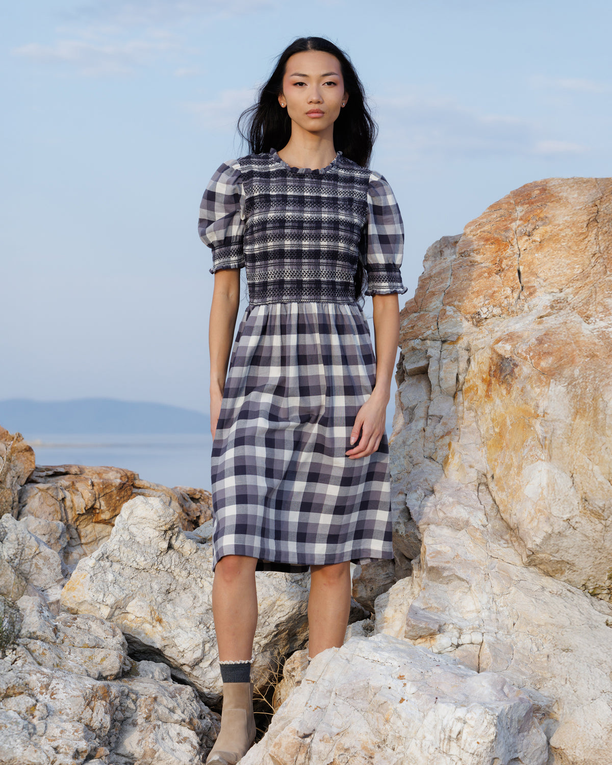 A model stands on rocky coastal terrain wearing a black and white gingham midi dress with puff sleeves and a smocked bodice.