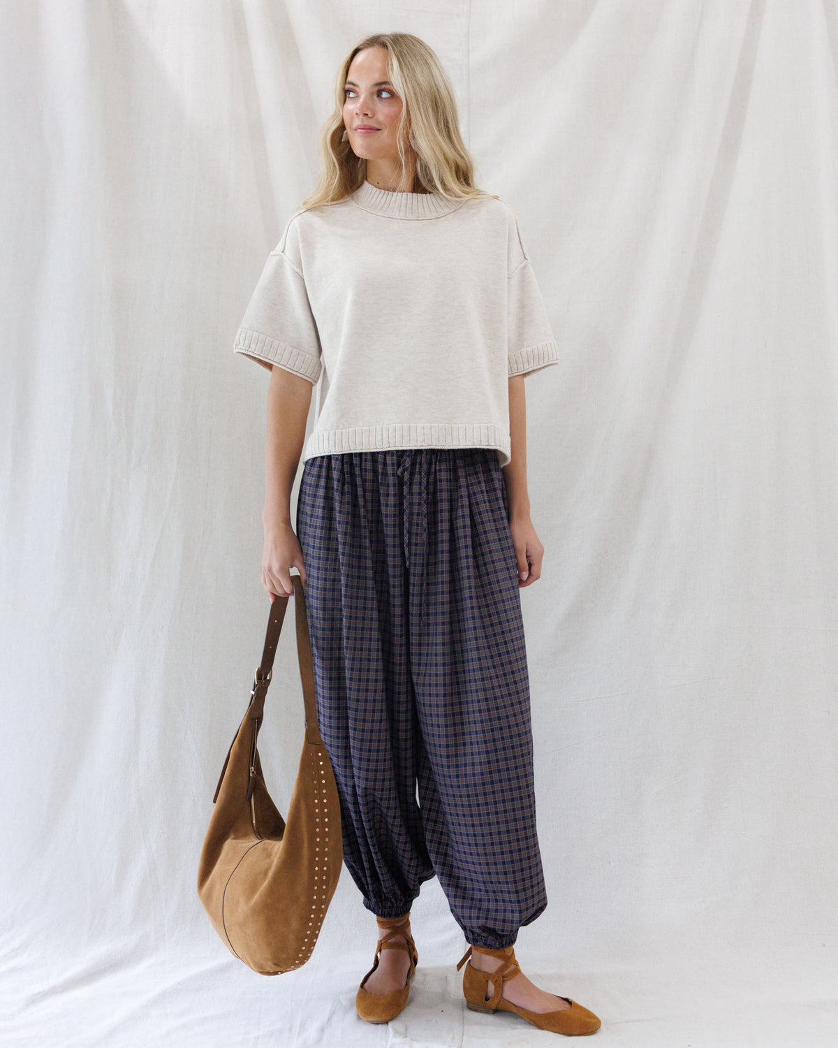 Front full-length view of the boxy beige short-sleeve knit sweater paired untucked with high-waisted navy and red plaid balloon pants.