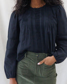Front close-up of a black long-sleeve cotton blouse featuring vertical pintuck pleats, delicate lace ladder inserts down the bodice, a ruffle neckband, and elasticized cuffs.