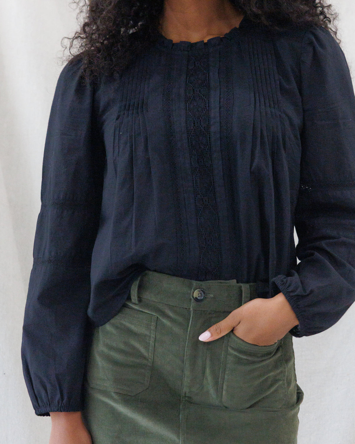 Front close-up of a black long-sleeve cotton blouse featuring vertical pintuck pleats, delicate lace ladder inserts down the bodice, a ruffle neckband, and elasticized cuffs.