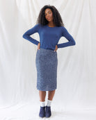 A woman modeling a blue long-sleeve crewneck top and a sparkly blue sequined pencil skirt, paired with white socks and navy blue boots, standing against a white fabric backdrop.